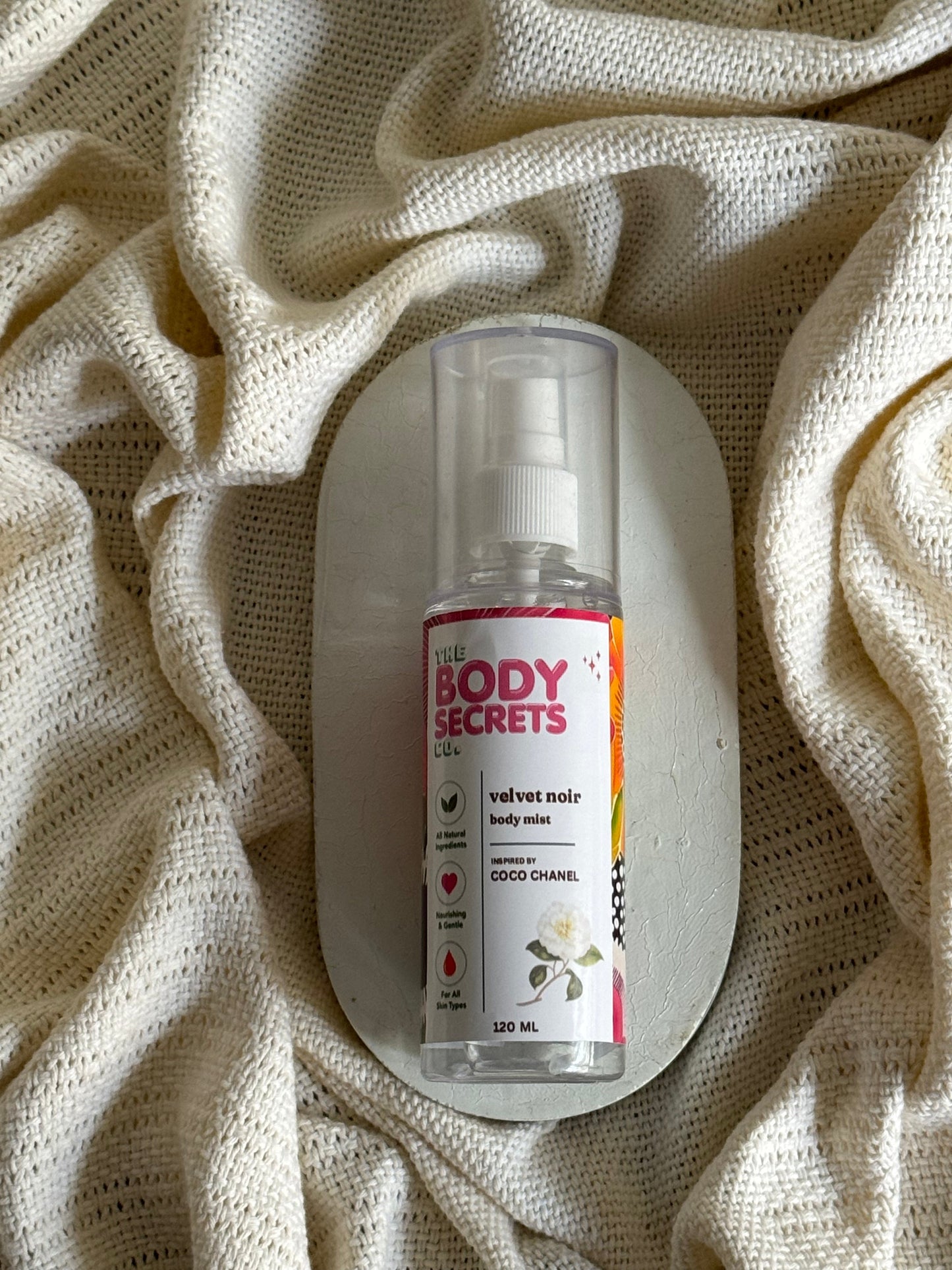 A clear plastic body spray bottle labeled 'BODY SECRETS' and 'GOOD CHAMILE' with a white pump, resting on a white oval dish over a textured cream knit blanket.